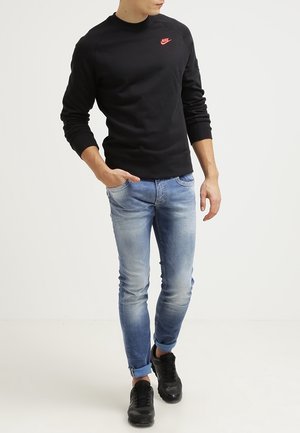 Sudadera negra de manga larga con el logo rojo de Nike, combinada con jeans desgastados de un azul claro y zapatillas negras. Modelo posicionado de manera casual.