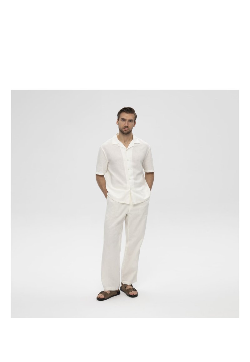 Man staat met handen in de zakken, draagt een wit kort mouw overhemd met knopen, witte broek en donkere sandalen op een effen grijze achtergrond.