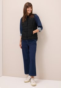 Donna sorridente che indossa un gilet trapuntato nero, una camicia di jeans con maniche arrotolate, pantaloni jeans blu scuro e sneaker bianche, in piedi contro un muro beige.