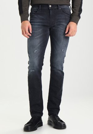 Mężczyzna noszący dopasowane, ciemnoniebieskie, postarzane dżinsy i czarne sznurowane buty, z rękami swobodnie opuszczonymi wzdłuż ciała, na neutralnym tle.