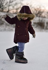 Vyno spalvos parka su kailiniu gobtuvu, dviem kišenėmis, sagomis ir pirštinėmis. Vaikas sniego batais stovi ant snieguoto žemės, demonstruodamas mėlynas džinsines kelnes.