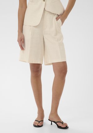 Žena v béžových plisovaných šortkách po kolená, sako v rovnakej farbe a čierne sandále na nízkom podpätku, stojaca s jednou rukou v vrecku.