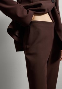 Gros plan sur une personne portant un pantalon de costume taillé marron foncé et un blazer assorti, la main glissée dans la poche du pantalon sur un fond uni.