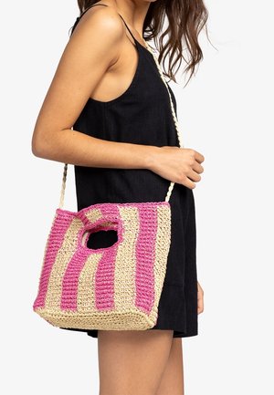 Mujer con vestido negro sin mangas que lleva un bolso de hombro tejido con rayas beige y rosa, con un agujero integrado para el asa.