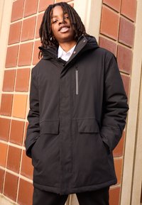 Sort vandtæt parka med hætte, lynlås foran, to store lommer og en lynlåslomme på brystet. Glat tekstur, minimalistisk design.