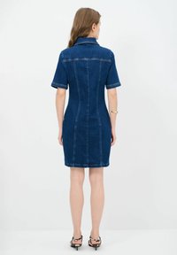 Denim-kjol med korta ärmar, åtsittande silhuett, krage, synliga sömnadsdetaljer och längd till mitten av låret. Mörkblå färg med lätt textur.