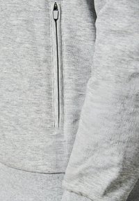 Zbliżenie na jasnoszary prążkowany materiał z pionową kieszenią na zamek i mankietem rękawa, prawdopodobnie część bluzy lub kurtki.