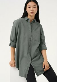 Pikad varrukad, rohelise oliivitooniga nööbitav särk, valmistatud pehmest kangast, millel on rinnatasku ja ülesrullitud varrukad, veidi kumer alläär.