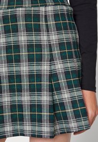 Grüne und marineblaue Karo-Minirock mit Falten. Verfügt über weiße und goldene Nähte, die das Karomuster kreuzen. Hergestellt aus strukturiertem Stoff.