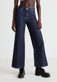 Jeans a gamba larga in denim scuro con cuciture bianche a contrasto e tasche, abbinati a scarpe con tacco nero. Texture liscia e vestibilità rilassata.