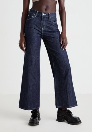 Wide-leg dark denim jeans with contrasting white stitching and pockets, paired with black platform shoes. Smooth texture and relaxed fit.