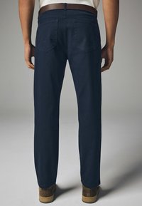 Marineblaue Jeans mit geradem Schnitt, ausgestattet mit zwei Gesäßtaschen und dezenten Nähten. Kombiniert mit braunen Schuhen.