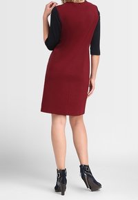 Abito aderente bordeaux con maniche nere a tre quarti, texture liscia, cerniera sul retro, abbinato a stivali alla caviglia blu con fibbie.