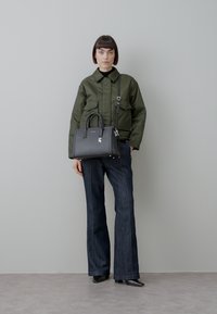 Svart skinnhåndveske med strukturert design, sølvfarget metallbeslag og håndtak på toppen. Modellen har på seg en mørkegrønn jakke og vidbente denim-bukser.