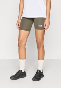 Βερμούδες ποδηλασίας σε ελαιόχρωμο πράσινο από ελαστικό ύφασμα, με λευκό λογότυπο. Συνδυάζονται με μαύρα αθλητικά παπούτσια και λευκές κάλτσες μεσαίου μήκους.