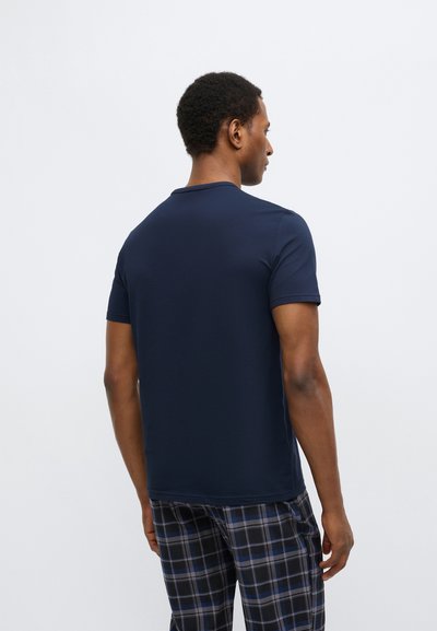 Tamsiai mėlynas trumpomis rankovėmis marškinėliai iš lygaus medvilninio audinio, apvalia iškirpte, matomi iš nugaros, derinami su languotomis pižamos kelnėmis.