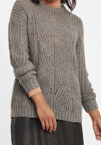 Donna che indossa un maglione grigio a maglia tessurizzata sopra un dolcevita beige, abbinato a una gonna di pelle nera, su uno sfondo semplice.