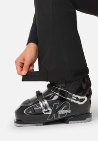 Musta laskettelumono kiiltävällä viimeistelyllä, hopeisilla metalliosilla, solkikiinnityksillä ja kontrastivärisellä logolla. Säädettävä istuvuus tarrakiinnitteisellä varresuojuksella.