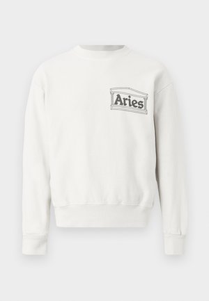 Muž stojaci s rukami v vreckách, oblečený v bielej košeli s dlhým rukávom s logom "Aries" a voľné tmavé nohavice s čiernymi topánkami, jednoduché pozadie.