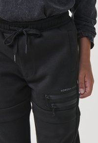 Detailný záber na čierne teplákové nohavice s elastickým pásom na šnúrku, dvojitými zipsovými bočnými vreckami a logom "Koroshi Brand" umiestneným na stehne, nosené s tmavým vrchom.
