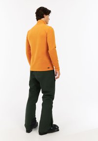 Oranžový fleece pulover s vysokým golierom, skombinovaný s tmavozelenými lyžiarskymi nohavicami. Čierne topánky s modrými akcentmi, pohľad zozadu.