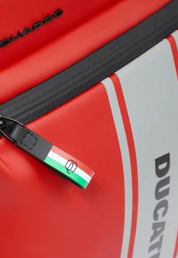 Bolsa de la marca Red Ducat con un acento gris, que presenta una cremallera prominente y una etiqueta de cuero negra con un diseño de bandera tricolor.