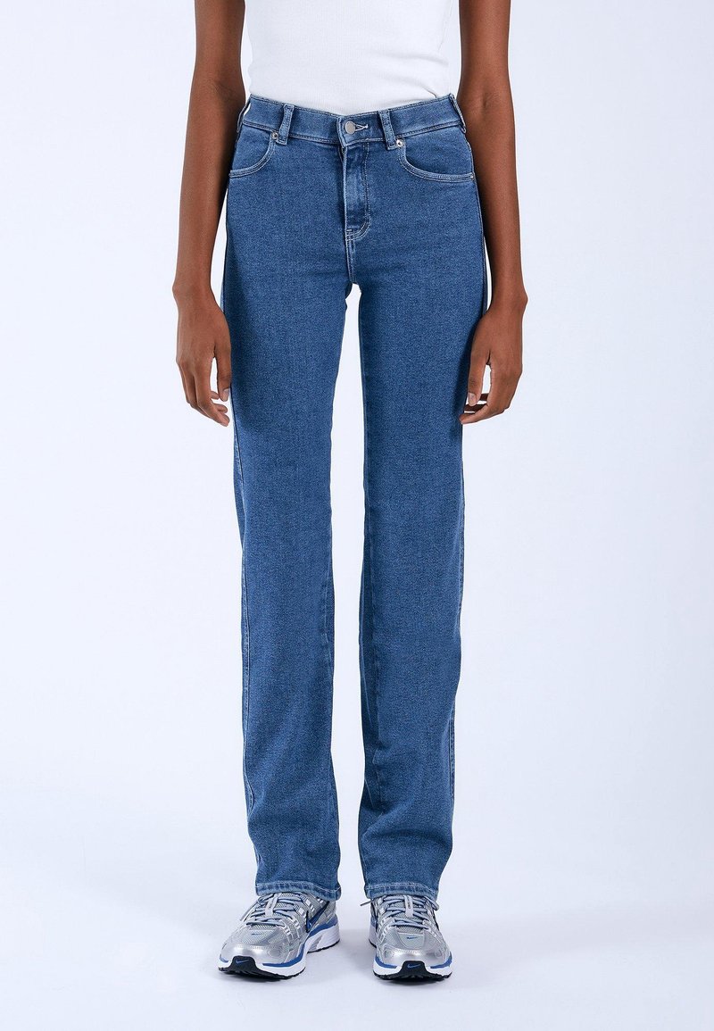 Jean en denim bleu avec une coupe droite, design à cinq poches et détails de couture discrets, associés à des chaussures de sport grises.