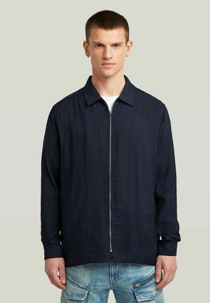 Marineblaue Zip-Jacke mit klassischem Kragen, aus leichtem Material und mit langen Ärmeln. Verfügt über ein minimalistisches Design und subtile Details.