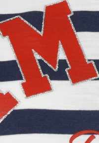 Roter Buchstabe "M" mit silbernem Rand auf weiß-blau gestreiftem Stoff, der eine weiche Textur und auffällige Designelemente aufweist.