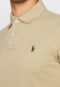 Beige pikétröja i texturerat tyg, med en grön broderad logotyp på vänster bröst och en klassisk krage med två knappar.