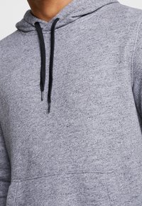 Grå hoodie med strukturerat tyg, justerbar svart dragsko och en ficka framtill, designad för komfort och praktisk användning.