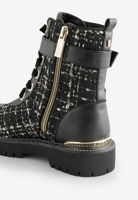 Stivaletto in pelle nera con tomaia in tessuto caratterizzata da un motivo intrecciato, zip laterale con finiture dorate e suola in gomma testurizzata.