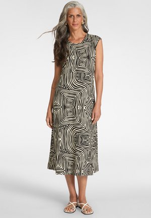 Mujer con cabello largo gris y negro que lleva un vestido midi sin mangas con un patrón geométrico en blanco y negro y sandalias blancas de tiras.