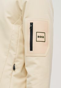 Manche de veste beige avec une poche rectangulaire zippée et un patch logo "BOSS" près du haut du bras.