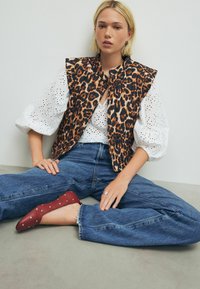 Een leopard print vest met een hoge kraag, gecombineerd met een witte blouse met oogjes. Blauwe jeans en bordeaux rode ballerina's met zilveren studs.