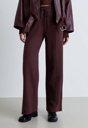 Personne portant un pantalon large taille haute marron foncé, des chaussures noires épaisses, un top en dentelle bordeaux et une veste en cuir bordeaux avec ceinture et détails zippés.