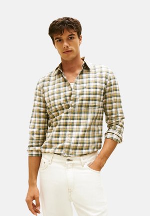 Giovane uomo con una camicia a quadri verde e gialla a maniche arrotolate e pantaloni bianchi, in piedi con una mano in tasca.