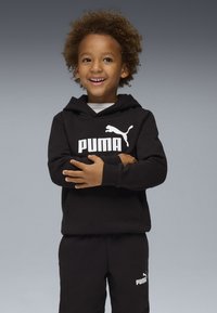 Haut à capuche noir et pantalon de jogging avec le logo blanc de Puma. Caractéristiques : coupe décontractée, poche kangourou et texture douce. L'enfant se tient avec les bras croisés.