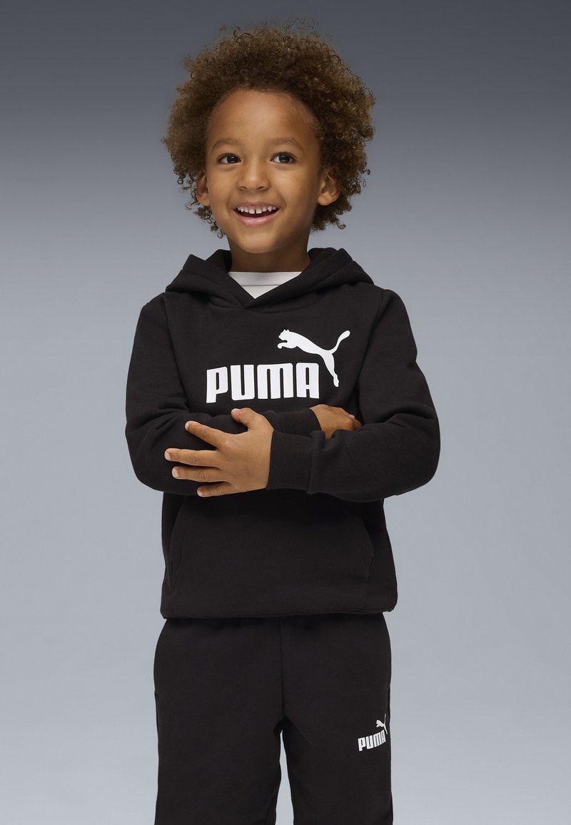 Schwarzer Hoodie und Sweatpants mit weißem Puma-Logo. Verfügt über eine entspannte Passform, eine Kängurutasche und eine weiche Textur. Kind steht mit verschränkten Armen.