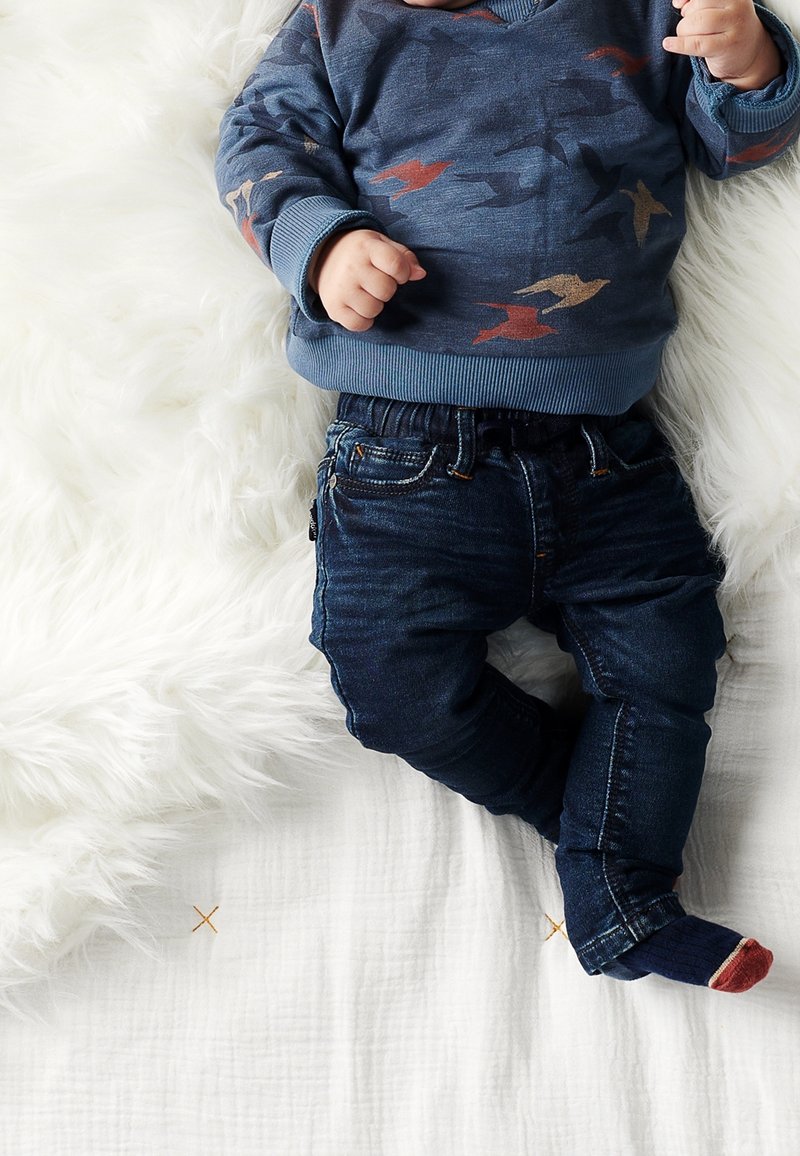 Bébé portant un pull bleu à motifs d'oiseaux, un jean foncé et des chaussettes dépareillées, allongé sur une literie blanche texturée avec une couverture blanche duveteuse.