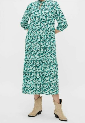 Grünes und weißes midikleid mit Blumenmuster und langen Ärmeln, kombiniert mit beigen Wildleder-Cowboystiefeln bis zur Mitte der Wade, an einer stehenden Person.