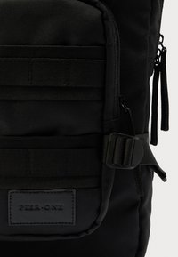 Sac à dos en tissu noir avec une texture lisse, plusieurs sangles, une fermeture éclair et un patch en cuir embossé du texte « PIER • ONE ».
