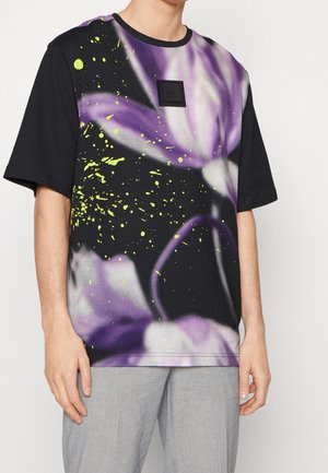Homme portant un T-shirt noir à manches courtes avec un grand motif de fleur violette et des éclaboussures jaunes, associé à un pantalon gris clair.