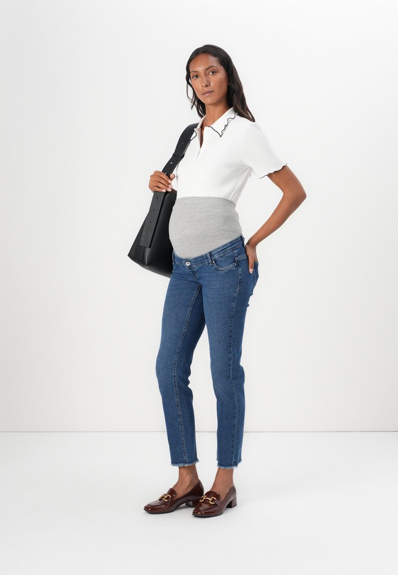 Weißes Poloshirt mit Rüschensaum, grauem Einsatz, blauen Slim-Fit-Jeans und braunen loafers. Das Model hält eine schwarze Umhängetasche.