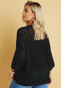 Schwarzer, gestrickter Pullover mit offener Webstruktur, überschnittenen Schultern und Dreiviertelärmeln. Getragen über blauen Jeans.