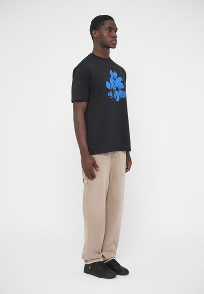 T-shirt noir avec un motif floral bleu, associé à un pantalon cargo beige et des baskets noires. Le mannequin est de profil sur un fond blanc.