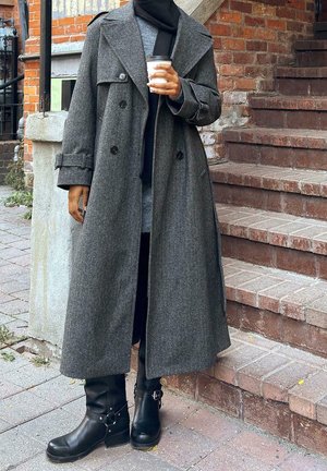 Persona con un abrigo largo gris, bufanda negra y botas negras, sosteniendo un vaso de café para llevar, de pie junto a unos escalones y una pared de ladrillo al aire libre.