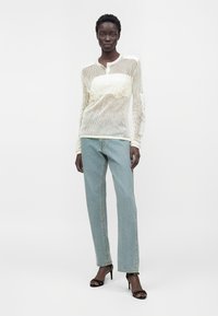 Femme portant un haut à manches longues en maille blanche, un jean bleu clair droit et des sandales noires à talons, debout devant un fond blanc uni.