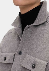 Szara wełniana koszula z złożonym kołnierzykiem, posiadająca dwie przednie kieszenie, czarne guziki oraz prążkowaną fakturę na kołnierzyku i mankietach.