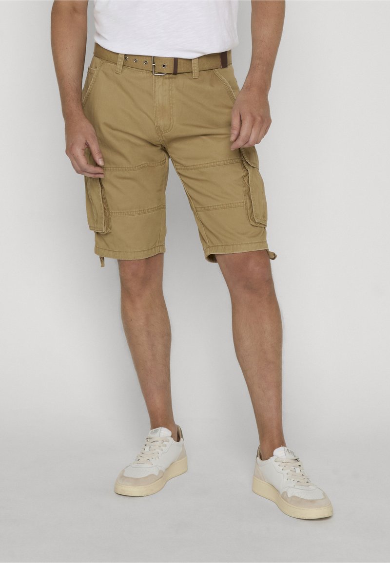 Homme portant un short cargo beige avec plusieurs poches, une ceinture marron, des baskets blanches et un t-shirt blanc, debout devant un fond uni.